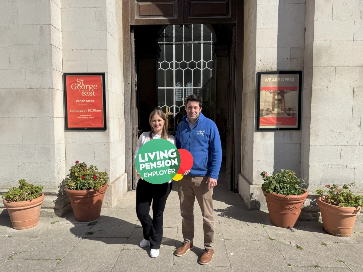 Two Clean for Good employees holding a Living Pension Employer logo foamboard standing outside a large stone doorway.