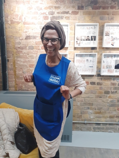 Clean for Good employee standing inside in front of a bare brick wall smiling and pointing to her branded cleaning overalls
