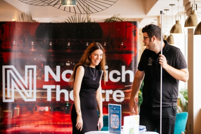 Two people in black stand in front of a red Norwich Theatre banner