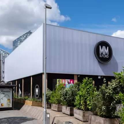 A grey building with a sign reading 'MM Ilford' outside is green bushes in wooden planters