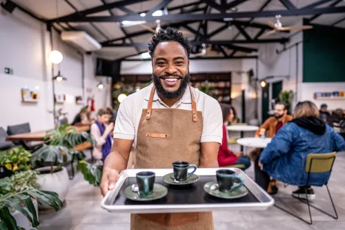 Waiter holds a tray of coffee while smiling at the camera. Cafe in the background.
