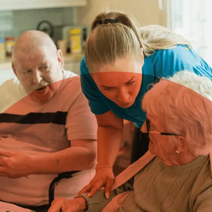 A caregiver helps an elderly woman in a wheelchair while an older man sits nearby. 